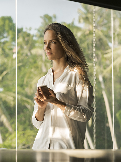 Donna dall'esprsssione serena opera con lo smartphone nella sua casa di design di fronte a un giardino lussureggiante.