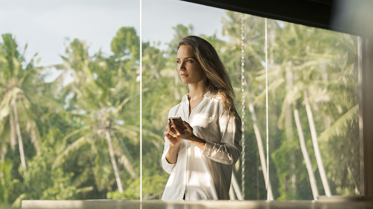 Donna dall'esprsssione serena opera con lo smartphone nella sua casa di design di fronte a un giardino lussureggiante.