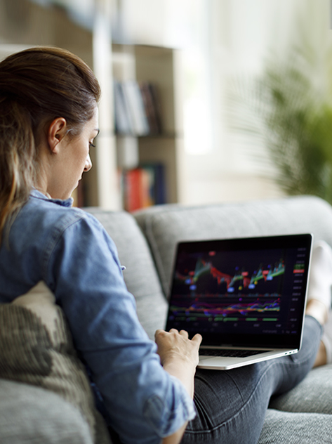 Giovane donna analizza grafici di andamento dei mercati sul proprio laptop, comodamente seduta sul divano di casa.