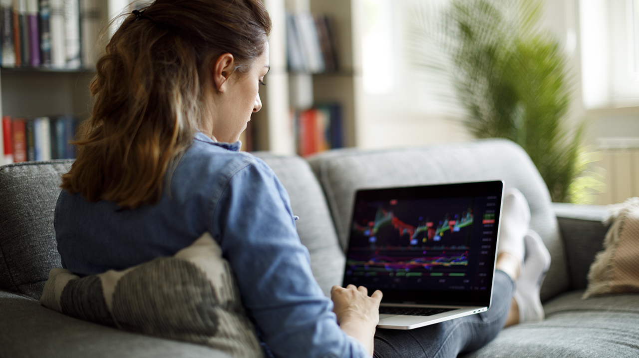 Giovane donna analizza grafici di andamento dei mercati sul proprio laptop, comodamente seduta sul divano di casa
