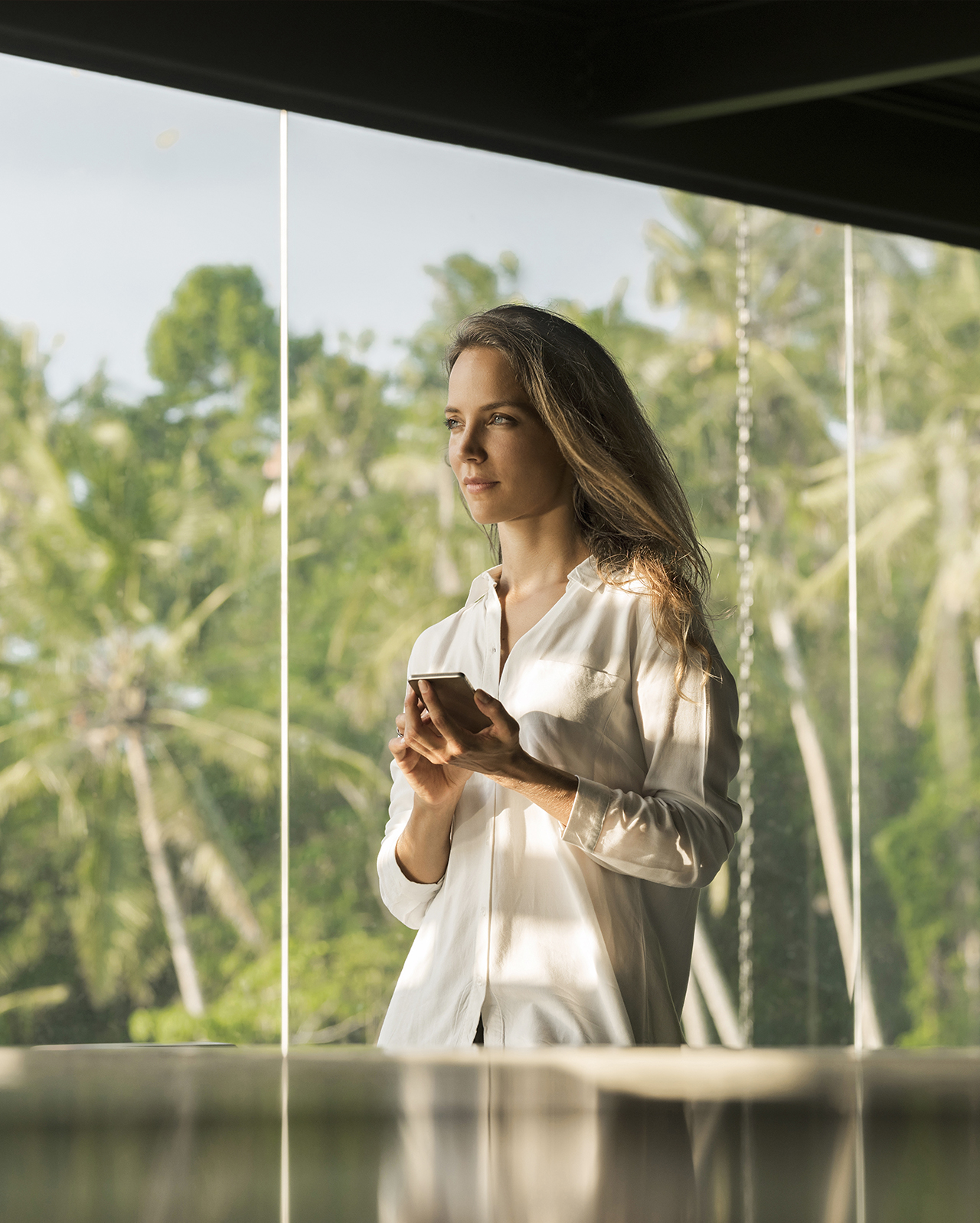 Donna dall'espressione serena opera con lo smartphone nella sua casa di design di fronte a un giardino lussureggiante.