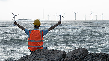 Un Ingegnere visto di spalle con due modelli di turbina eolica in mano, osserva in lontananza delle pale eoliche sul mare.