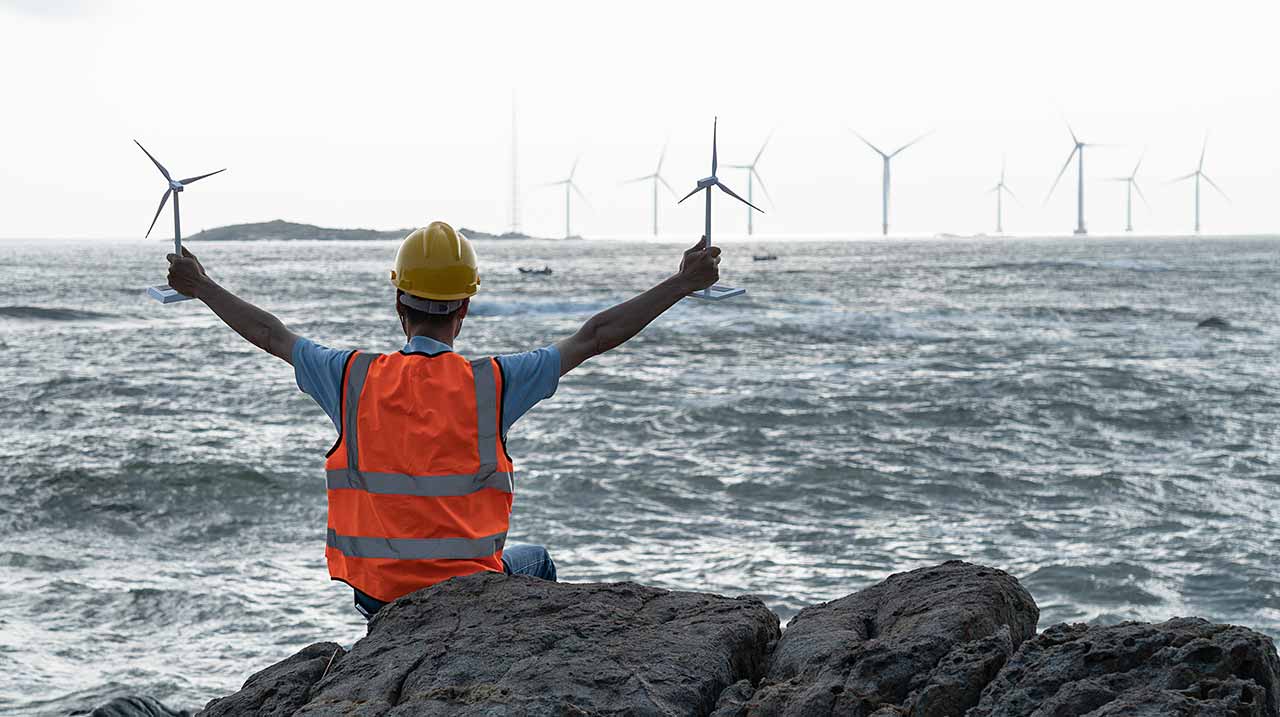 Ingegnere visto di spalle con due modelli di turbina eolica in mano, osserva in lontananza delle pale eoliche sul mare.