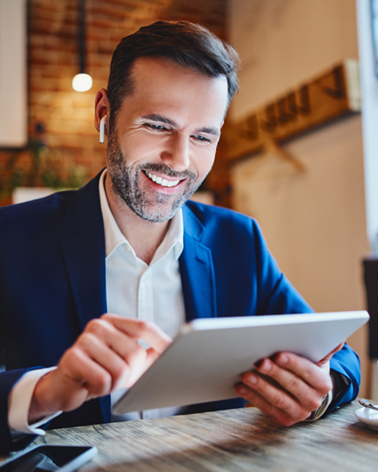 Uomo d'affari seduto al bar consulta il tablet e accede a contenuti multimediali con cuffie wireless.