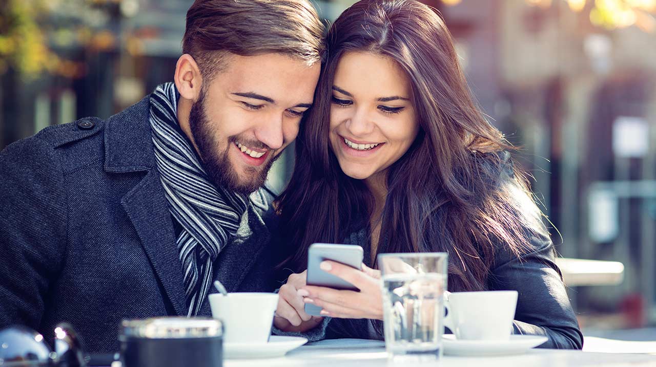 Una bella e giovane coppia di ragazzi navigano dal proprio smartphone in un caffè e sorridono.